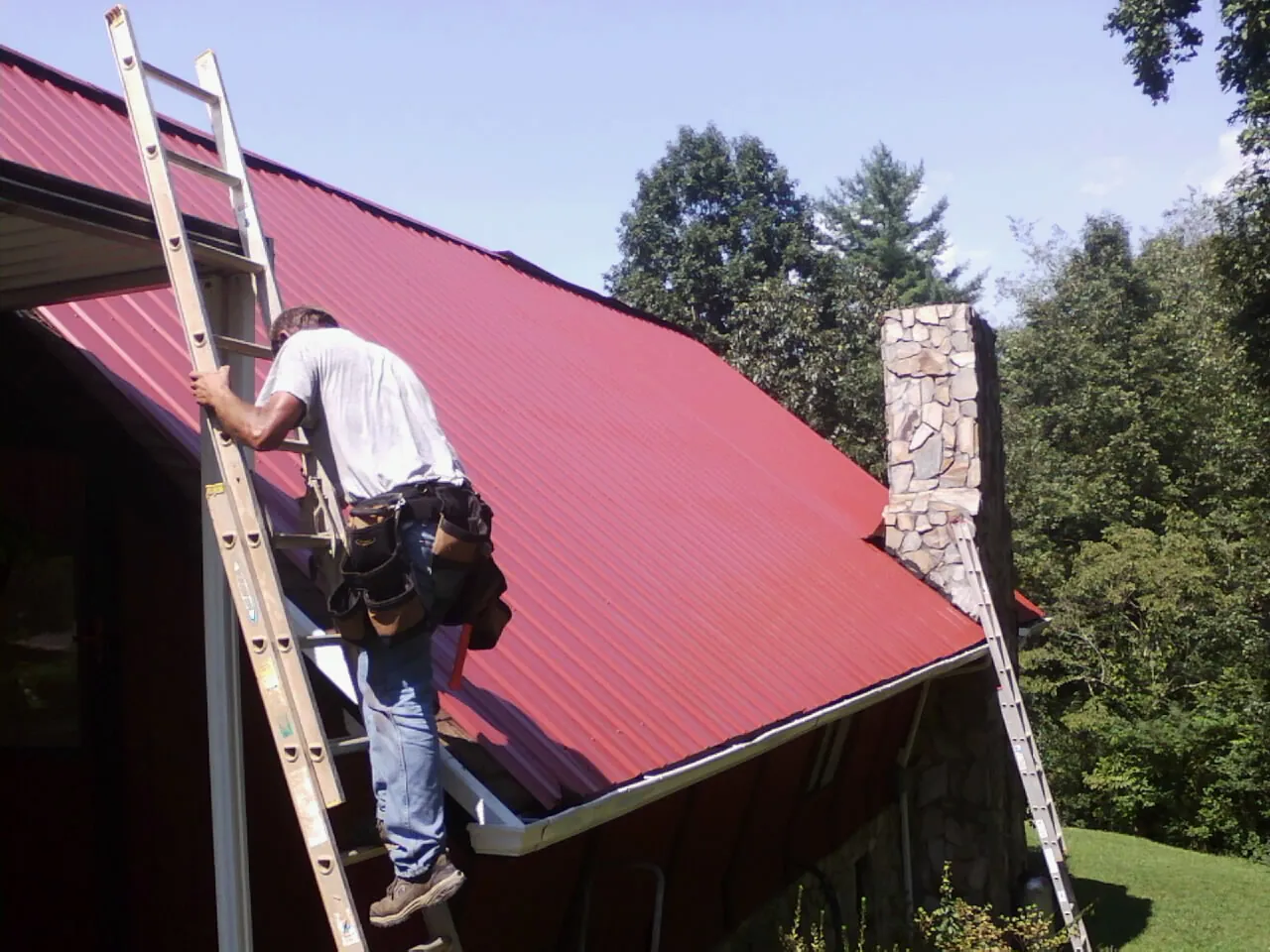 Roofer on a ladder beside a red metal roof and stone chimney — Soffit & Fascia Repair by Roofing Brothers in Myrtle